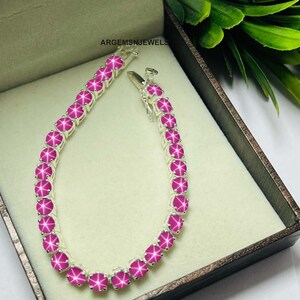 May include: A silver bracelet with a row of pink star-shaped gemstones. The bracelet is displayed in a brown velvet box.