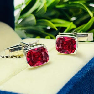 May include: A pair of silver cufflinks with large, square, deep red gemstones. The cufflinks are set against a light cream-colored surface, with green foliage in the background. The text "ARGEMSNJEWELS" is visible.