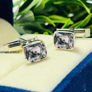 Puede incluir: Gemelos de plata con piedras cuadradas de ágata musgosa translúcida. Las piedras tienen un patrón natural verde en forma de helecho. Los gemelos se muestran en una caja azul con un interior crema, con follaje verde de fondo.