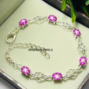 May include: A silver bracelet with a chain link design and six pink star sapphire gemstones. The bracelet has a lobster clasp closure. ARGEMSN JEWELS is printed on the bracelet.