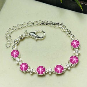 May include: A silver bracelet with a chain and clasp, featuring five pink star ruby gemstones set in silver with small white gemstones around each ruby. The bracelet has the text "ARGEMSNJEWELS" on the chain.