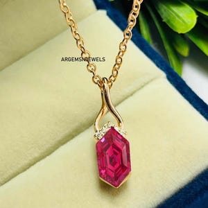 Peut inclure: Un collier doré avec un pendentif orné d'une pierre précieuse rose foncé facettée. Le pendentif est de forme géométrique avec un sertissage doré et de petites pierres d'accent claires. La chaîne est de style maillon délicat. Le texte "ARGEMSNJEWELS" est visible.