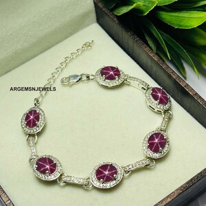 Puede incluir: Un brazalete de plata con cinco piedras preciosas ovaladas de rubí rojo rodeadas de piedras preciosas más pequeñas y transparentes. El brazalete tiene un cierre de langosta.