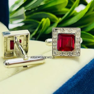 May include: Silver cufflinks with a square design. Each cufflink features a large, square, deep red gemstone at the center, surrounded by smaller, clear stones. The cufflinks are set against a backdrop of green foliage and a blue velvet surface.