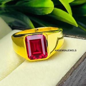 Op de afbeelding: Een goudkleurige ring met een grote, rechthoekige, robijnrode edelsteen. De ring is gezet in een dikke, gepolijste gouden band. De ring wordt gepresenteerd op een lichtgekleurd oppervlak met groen gebladerte op de achtergrond. De tekst "ARGEMSNJEWELS" is zichtbaar.