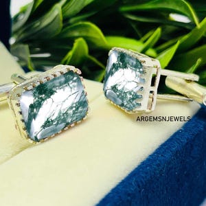 Puede incluir: Un par de gemelos plateados con piedras rectangulares de ágata musgosa. Las piedras presentan un patrón verde y blanco. Los gemelos se exhiben sobre una superficie amarillo claro, con follaje verde de fondo. El texto "ARGEMSNJEWELS" es visible.