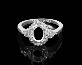 May include: A silver ring with a large oval-shaped setting surrounded by smaller diamonds. The ring is set on a black background.  ARGEMSNJEWELS is printed on the ring.