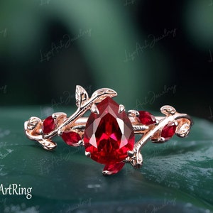Könnte beinhalten: Ein Ring mit einem birnenförmigen Rubin-Edelstein, der in Roségold gefasst ist und ein zartes Rankendesign mit kleinen Rubin-Akzenten aufweist. Der Ring liegt auf einem grünen Hintergrund.