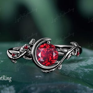 May include: A silver ring with a large, round, red gemstone. The ring has a crescent moon design with leaf and vine details and smaller red gemstones. The text "LagArtRing" is visible.