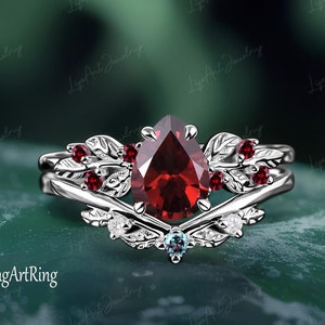 Puede incluir: Un anillo de plata con una piedra roja en forma de pera y acentos en forma de hoja. El anillo está engastado con piedras rojas más pequeñas y una sola piedra azul.