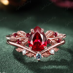 Puede incluir: Un anillo de oro rosa con una gran piedra central roja en forma de pera, acentuada por piedras rojas redondas más pequeñas y piedras transparentes engastadas en una banda con motivos de hojas. El metal tiene un tono dorado rosado cálido.