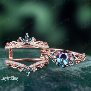 Könnte beinhalten: Ein Ring aus Roségold, besetzt mit einem großen, runden, blauen Edelstein, der von kleineren blauen Edelsteinen umgeben ist. Der Ring ist mit filigranen Blattmotiven verziert. Ein passender Ring aus Roségold mit einem Kronendesign und kleineren Edelsteinen ist ebenfalls abgebildet.