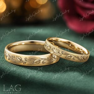 May include: Two gold wedding bands with a floral leaf design. The rings are set against a dark green background. The rings have a brushed finish and are engraved with leaves and vines.