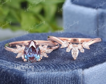 May include: A rose gold ring set featuring a pear-shaped, color-changing gemstone with surrounding marquise-cut stones. The set includes a matching curved band with additional accent stones. The rings are displayed on a blue velvet surface.