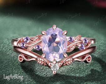 Puede incluir: Un anillo de oro rosa con una gran piedra central de corte ovalado en color morado claro, acentuado por piedras moradas redondas más pequeñas y detalles metálicos en forma de hoja a lo largo de la banda. Incluye una banda en forma de V.