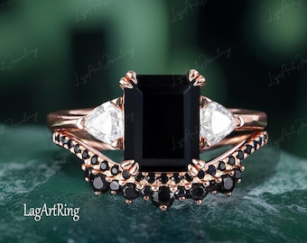 May include: A rose gold engagement ring set with a black emerald cut gemstone and two pear-shaped side stones. The band is accented with black gemstones.