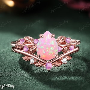 Pode incluir: Um anel de ouro rosa com uma pedra central de opala rosa em forma de pera. A banda é desenhada com motivos de folhas intrincados e acentuada com pequenas pedras redondas cor-de-rosa e pedras claras. O metal tem um acabamento polido, destacando o trabalho botânico detalhado em torno da opala central.