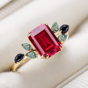 Puede incluir: Un anillo de oro con una gran piedra preciosa roja de talla esmeralda rodeada de pequeñas piedras preciosas verdes y negras. El anillo presenta un delicado diseño de hojas.