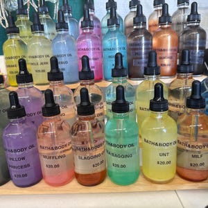 May include: A display of various colorful bath and body oils in clear glass bottles with black droppers. Each bottle has a label with a unique name and price. The oils are arranged on a wooden shelf.