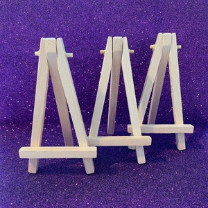 Puede incluir: Tres pequeños caballetes de madera natural, dispuestos en fila sobre un fondo morado brillante. Los caballetes son todos del mismo tamaño y forma, y están todos en foco.
