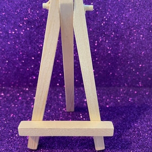 Könnte beinhalten: Ein kleiner, unfertiger Holzstaffelei mit einem dreibeinigen Design. Die Staffelei besteht aus hellfarbenem Holz und steht vor einem glitzernden, lila Hintergrund. Die Staffelei ist für die Aufnahme einer kleinen Leinwand oder eines Kunstwerks konzipiert.