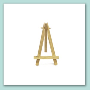 May include: A small, natural wood easel with a three-legged design. The miniature easel is made of light-colored wood and is suitable for displaying small artwork or photos. The easel is approximately 12 inches tall.