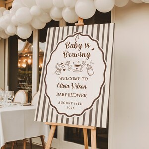 Può includere: Un cartello rettangolare su un cavalletto in legno con strisce verticali marroni e una cornice bianca smerlata. Il testo recita Baby is Brewing, Welcome to Olivia Wilson Baby Shower, August 24th 2026, con illustrazioni di caffettiera e biberon.