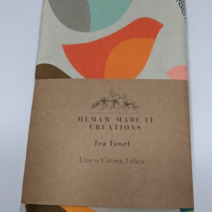 Pu&ograve; includere: Un asciugamano da t&egrave; con un motivo geometrico di forme arancioni, gialle, marroni e turchesi su sfondo bianco. L'asciugamano ha un'etichetta marrone su cui &egrave; scritto "Memaw Made It Creations Tea Towel Linen/Cotton fabric".