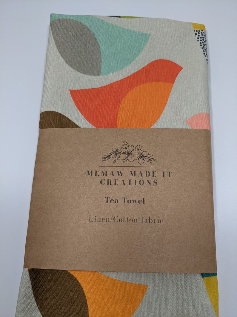 Pu&ograve; includere: Un asciugamano da t&egrave; con un motivo geometrico di forme arancioni, gialle, marroni e turchesi su sfondo bianco. L'asciugamano ha un'etichetta marrone su cui &egrave; scritto "Memaw Made It Creations Tea Towel Linen/Cotton fabric".