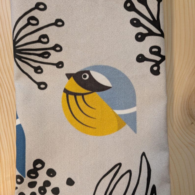 May include: A gray fabric with a stylized blue and yellow bird design. The bird has a black eye and a black beak. The background is a light gray with black floral designs.
