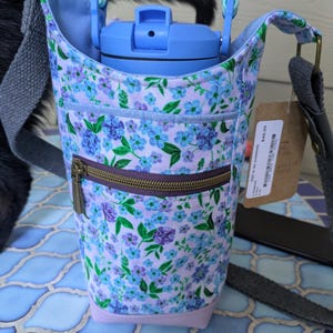 May include: A fabric water bottle carrier featuring a light purple background with a blue and green floral print. It includes a front zippered pocket, a light blue plastic water bottle, a braided blue handle, and a gray adjustable shoulder strap.