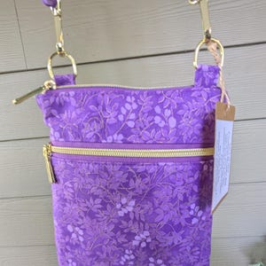 May include: A purple crossbody bag featuring a tonal leaf and floral pattern with gold-toned metal zippers and hardware. The bag includes an adjustable fabric strap attached with gold clips and a rectangular paper tag hanging from the side ring.