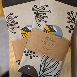 May include: Two tea towels with a black border and a light gray background. One towel features a black and white stylized bird with a yellow breast and a blue head. The other towel features a black and white stylized bird with a red breast. Both towels have a brown label that reads "Memaw Made It Creations Tea Towel Linen/Cotton Fabric".