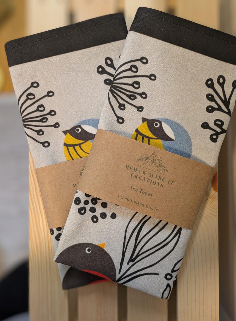 May include: Two tea towels with a black border and a light gray background. One towel features a black and white stylized bird with a yellow breast and a blue head. The other towel features a black and white stylized bird with a red breast. Both towels have a brown label that reads "Memaw Made It Creations Tea Towel Linen/Cotton Fabric".