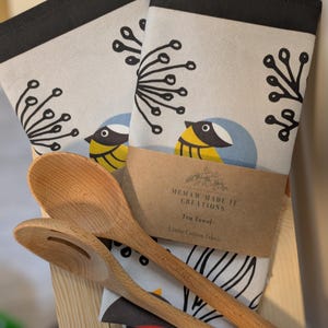 May include: Two tea towels with a black border and a white background featuring a stylized bird design with black, yellow, blue, and red accents. The text "MEMAW MADE IT CREATIONS" and "Tea Towel Linen/Cotton fabric" is printed on a brown tag attached to the towels.
