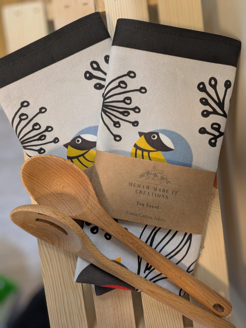 May include: Two tea towels with a black border and a white background featuring a stylized bird design with black, yellow, blue, and red accents. The text "MEMAW MADE IT CREATIONS" and "Tea Towel Linen/Cotton fabric" is printed on a brown tag attached to the towels.