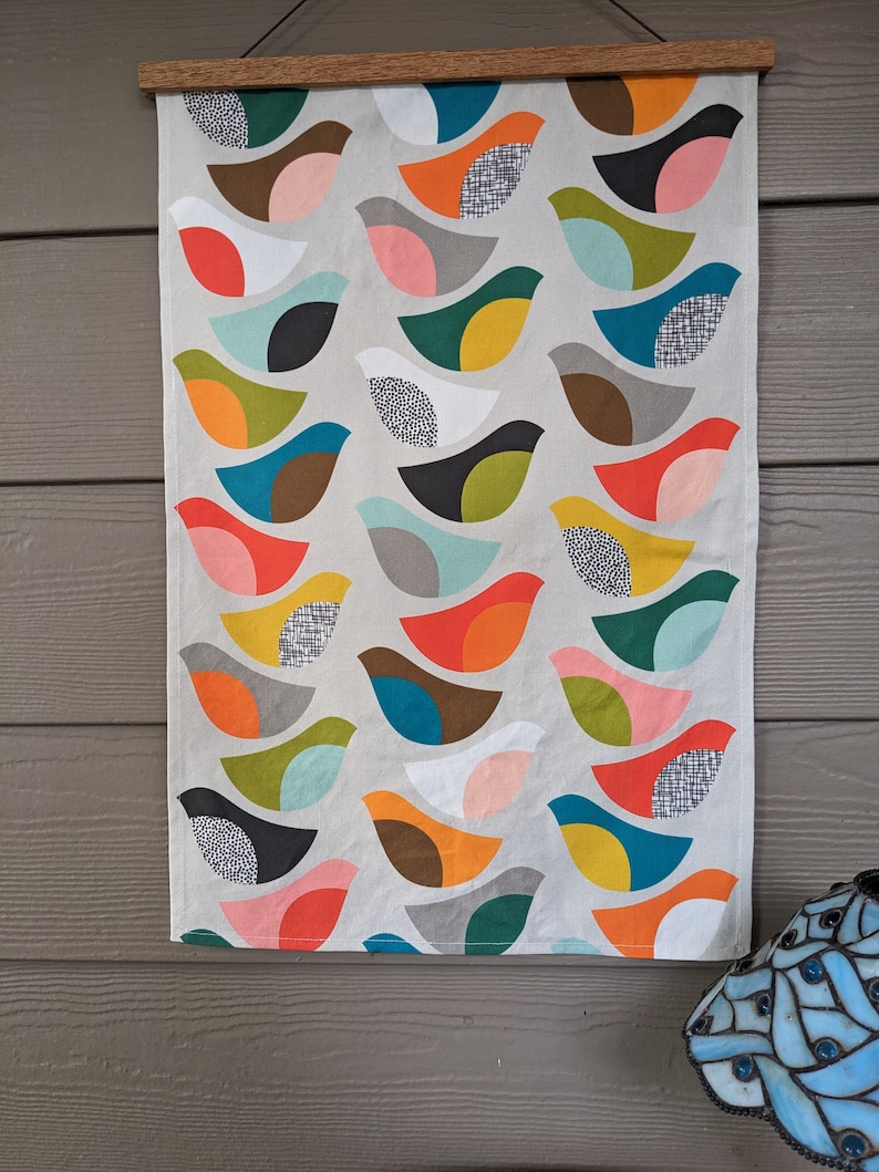 Pu&ograve; includere: Un arazzo da parete in tessuto bianco con un motivo ripetitivo di uccelli stilizzati colorati. Gli uccelli sono composti da forme geometriche in tonalit&agrave; di rosso, arancione, giallo, verde, blu, marrone e nero.