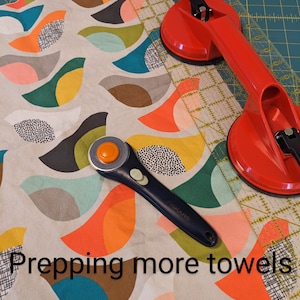 Pu&ograve; includere: Primo piano di un tessuto con un motivo geometrico colorato. Un taglierino rotativo, un righello e due ventose sono sopra il tessuto. Il testo "Prepping more towels" &egrave; visibile nella parte inferiore dell'immagine.