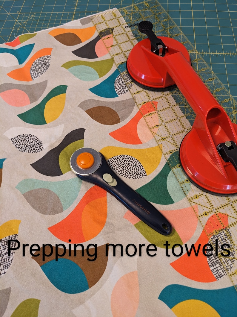 Pu&ograve; includere: Primo piano di un tessuto con un motivo geometrico colorato. Un taglierino rotativo, un righello e due ventose sono sopra il tessuto. Il testo "Prepping more towels" &egrave; visibile nella parte inferiore dell'immagine.