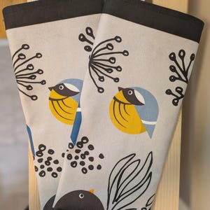 May include: Two beige tea towels with black borders feature a stylized design of birds, leaves, and berries. The birds are yellow, blue, and black. The leaves are black and white. The berries are black.