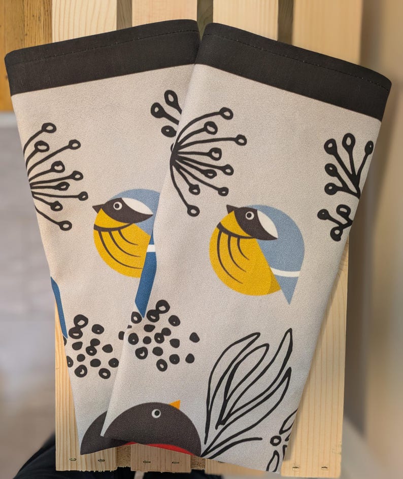 May include: Two beige tea towels with black borders feature a stylized design of birds, leaves, and berries. The birds are yellow, blue, and black. The leaves are black and white. The berries are black.