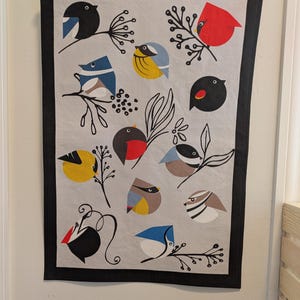 May include: A white kitchen towel with a black border features a colorful pattern of various birds with black branches and berries. The birds are in shades of blue, yellow, red, and black.