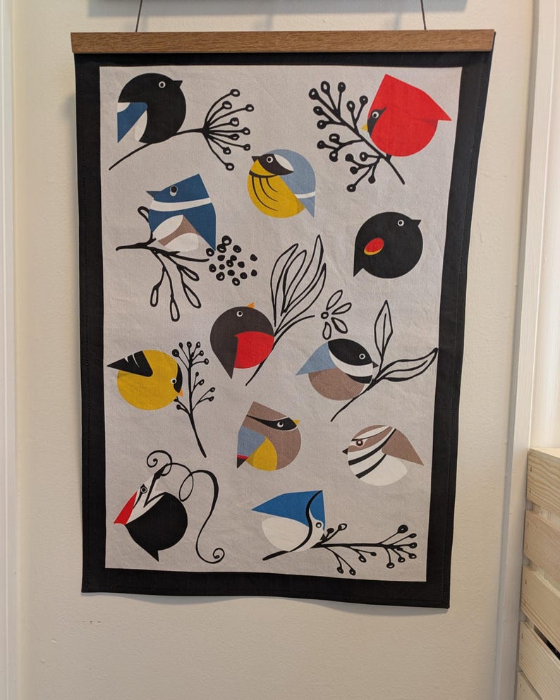 May include: A white kitchen towel with a black border features a colorful pattern of various birds with black branches and berries. The birds are in shades of blue, yellow, red, and black.