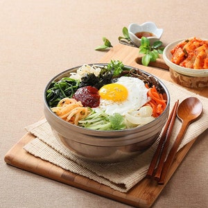 May include: A stainless steel bowl filled with a Korean bibimbap dish. The bowl contains rice, vegetables, a fried egg, and a red chili paste. The bowl is on a wooden cutting board with a small bowl of kimchi and chopsticks.