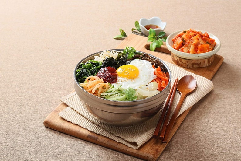 May include: A stainless steel bowl filled with a Korean bibimbap dish. The bowl contains rice, vegetables, a fried egg, and a red chili paste. The bowl is on a wooden cutting board with a small bowl of kimchi and chopsticks.