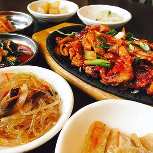 May include: A table setting with a variety of Korean dishes, including stir-fried meat, glass noodles, and various side dishes. The dishes are served in white bowls on a dark wooden table.