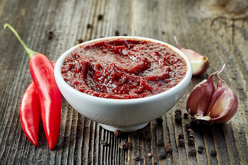 May include: A white bowl filled with a red chili sauce, surrounded by red chili peppers, garlic cloves, and black peppercorns on a wooden surface.