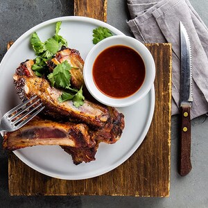 May include: A white plate with three pieces of barbecued pork ribs, a small white bowl with red barbecue sauce, and a sprig of cilantro.