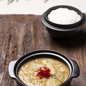 May include: A black bowl filled with a creamy soup topped with red peppers and green onions. The soup is served with a side of white rice in a black bowl.