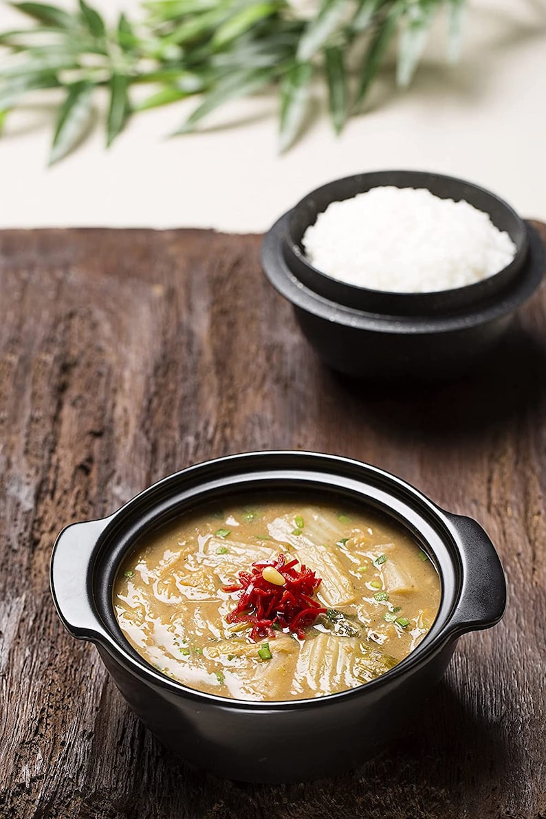 May include: A black bowl filled with a creamy soup topped with red peppers and green onions. The soup is served with a side of white rice in a black bowl.